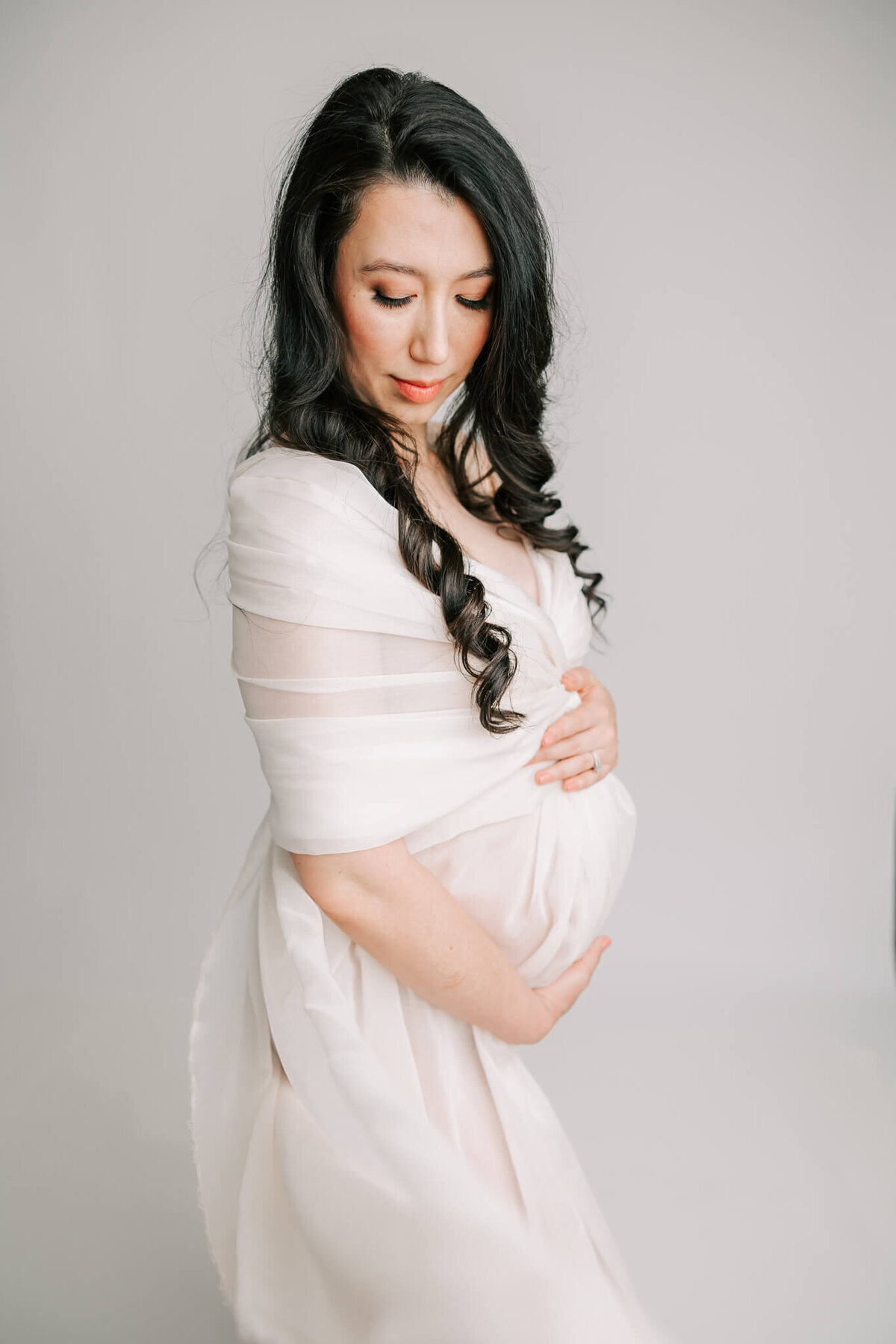 fine art maternity portrait of a woman in white silk. She had black hair and is holding her belly
