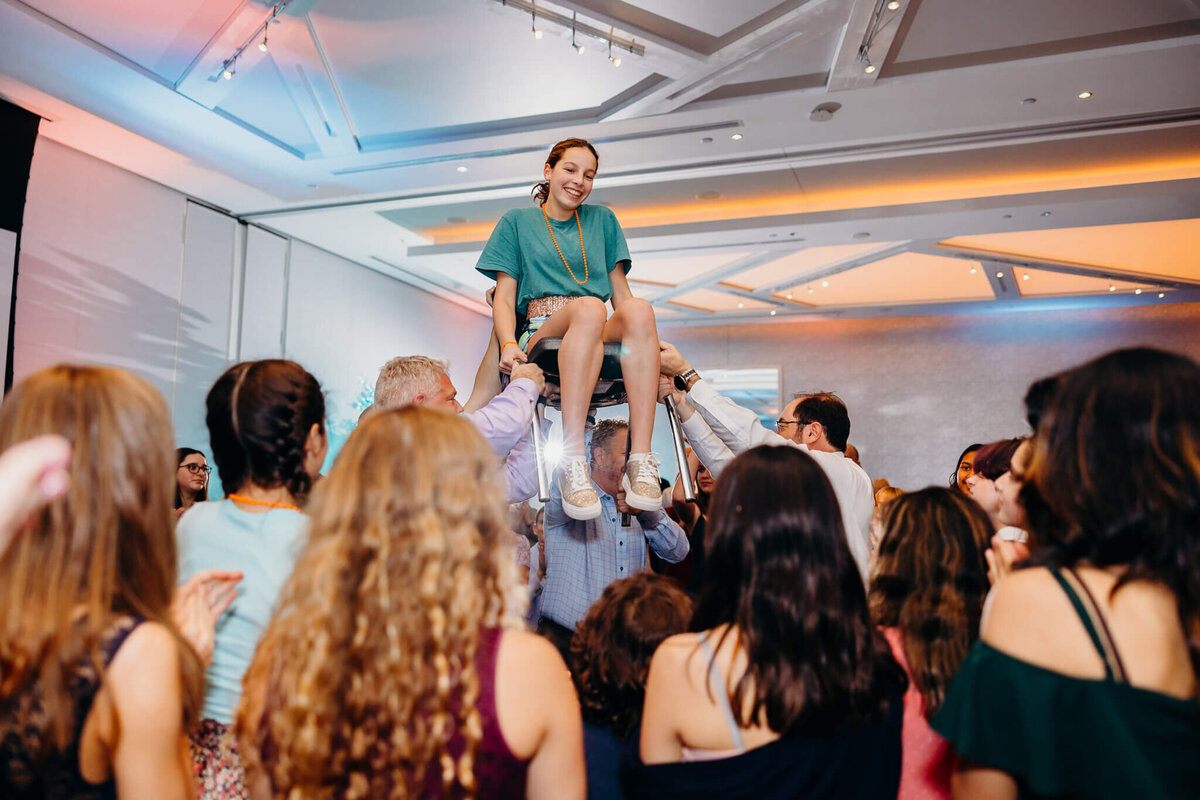 A teenage girl in a blue dress smiles while on the chair lifted up on the dance floor