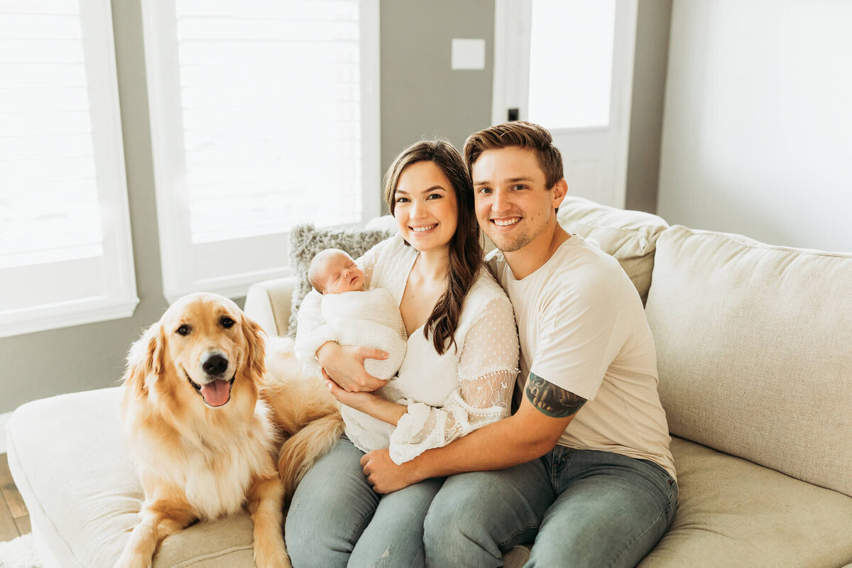 Family of three and their dog embracing the newest baby to join the family, while sitting on the couch together.
