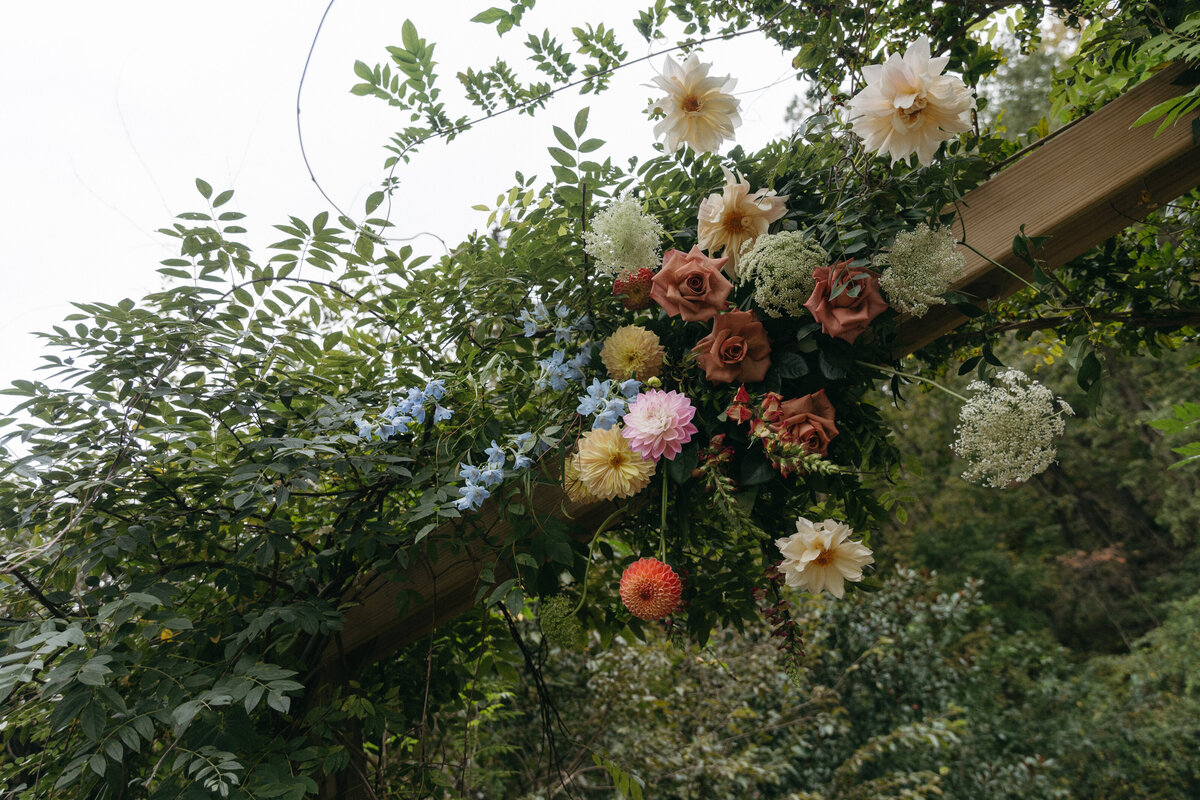 Close-up of a lush floral arch overflowing with dahlias, garden roses, delphinium, and queen anne’s lace, designed in a natural climbing style at The Nest outdoor ceremony in Northwest Arkansas.