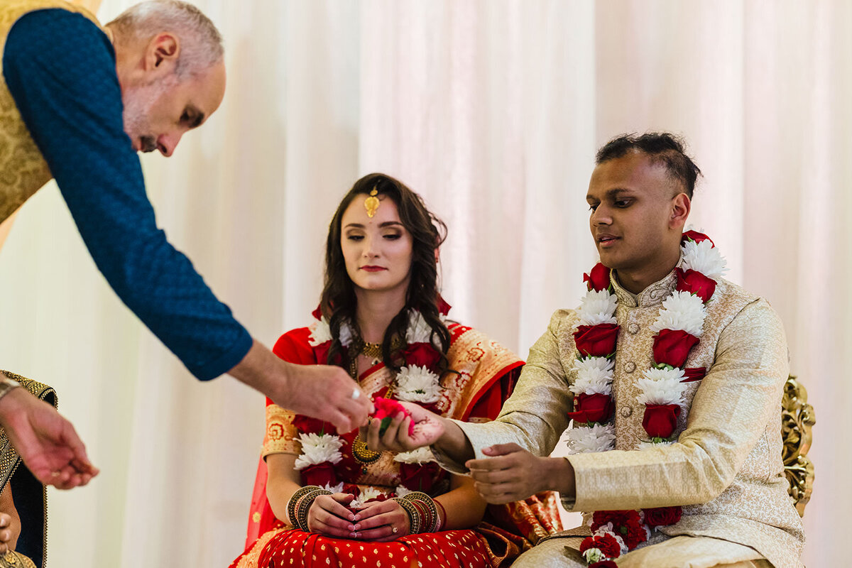 A man reaches out to give a ceremonial object to a bride and groom seated in traditional Indian wedding attire during a ritual.