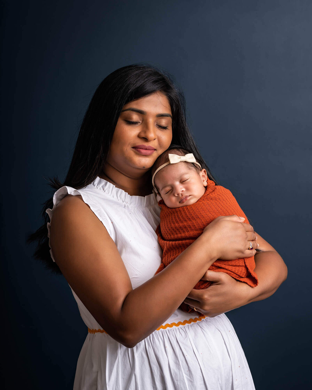 A mama in a white dress holds her newborn baby girl swaddled in a n orange blanket