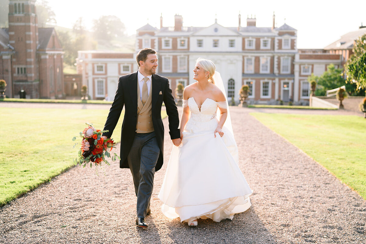 bride and groom walking towards camera at golden hour with hawkstone hall in the bckground of the photo