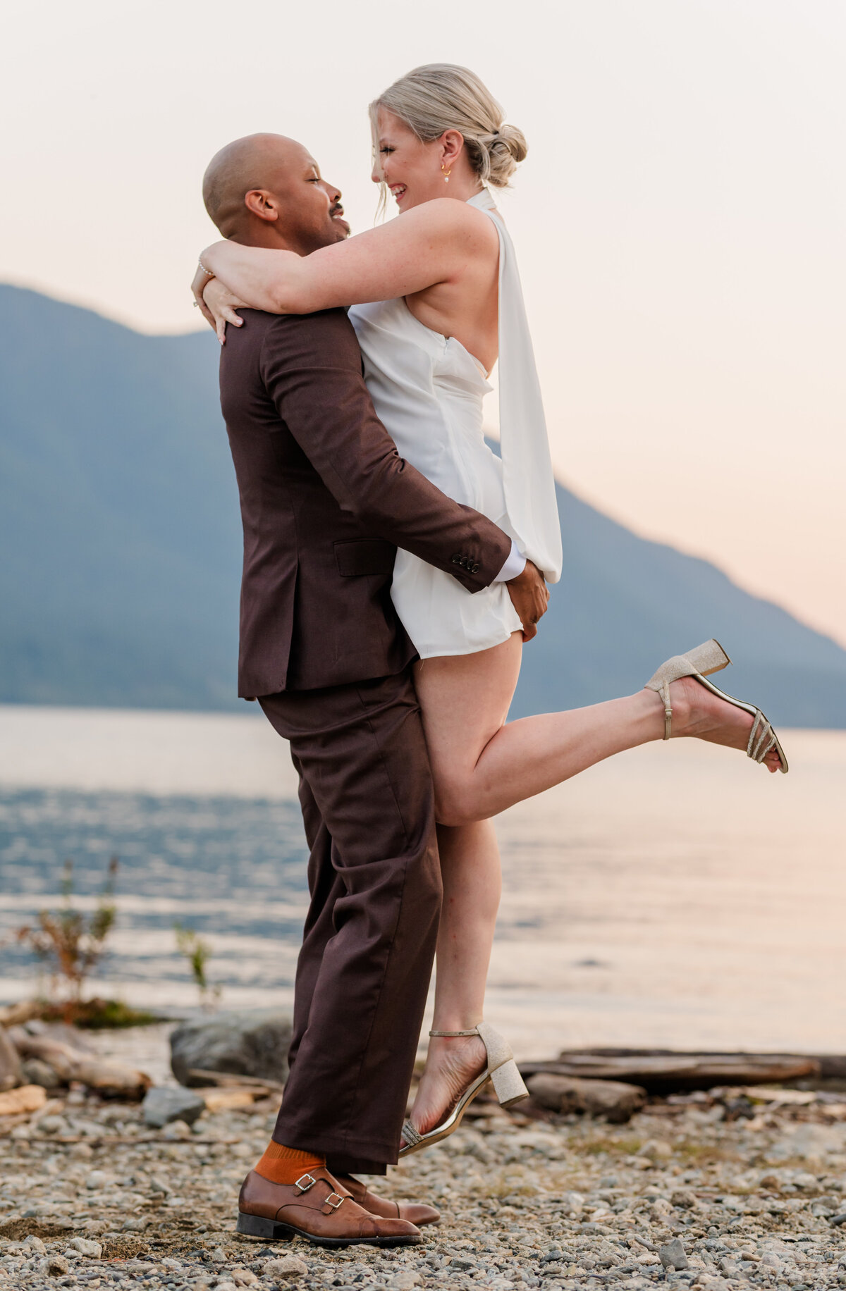 Guy picks up and the girls one leg popped out they are both looking at each. They are dressed up in brown and white attire with a lake and mountains behind them.