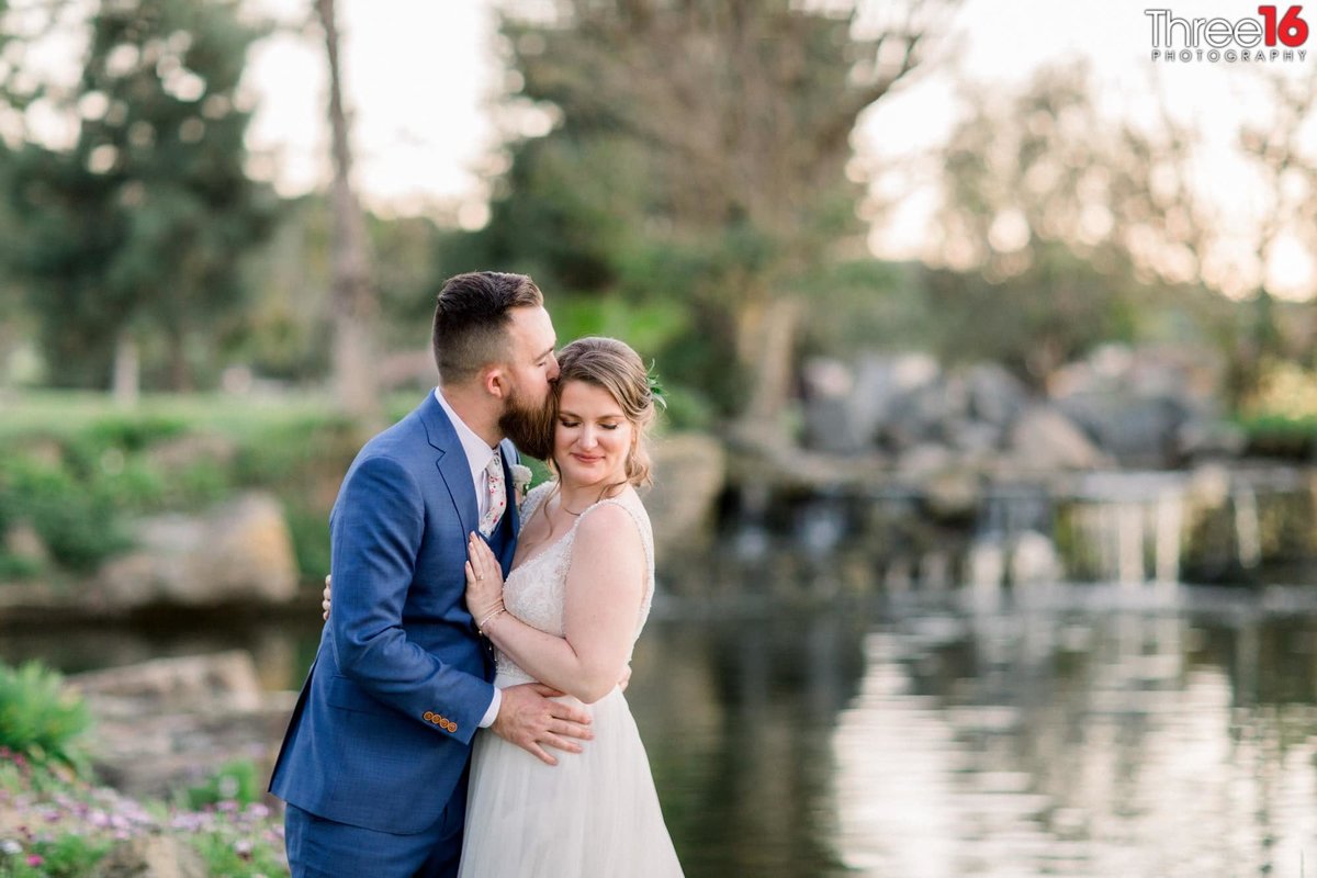 Groom kisses his Bride on the side of her head near the pond