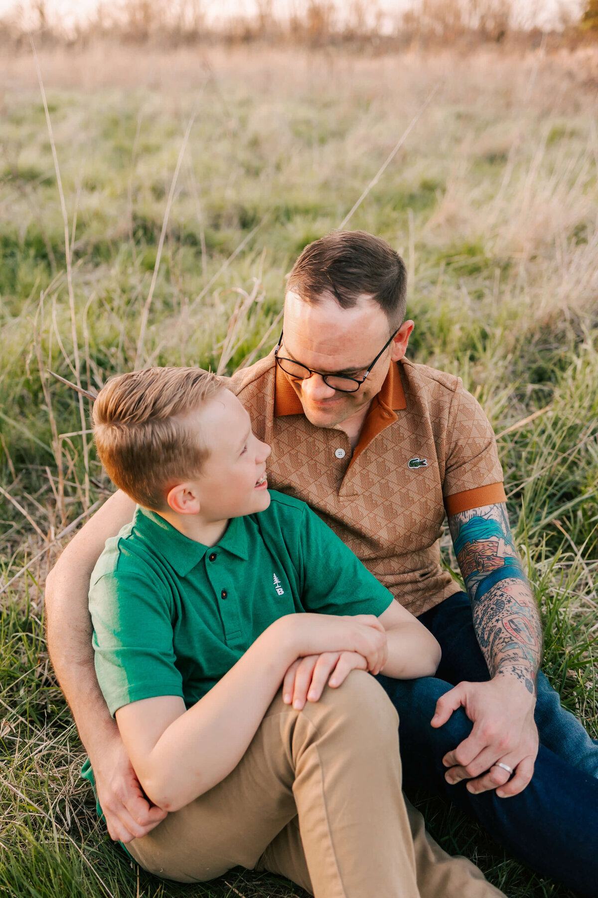 A father and son sitting together  in a field looking at each other and smiling