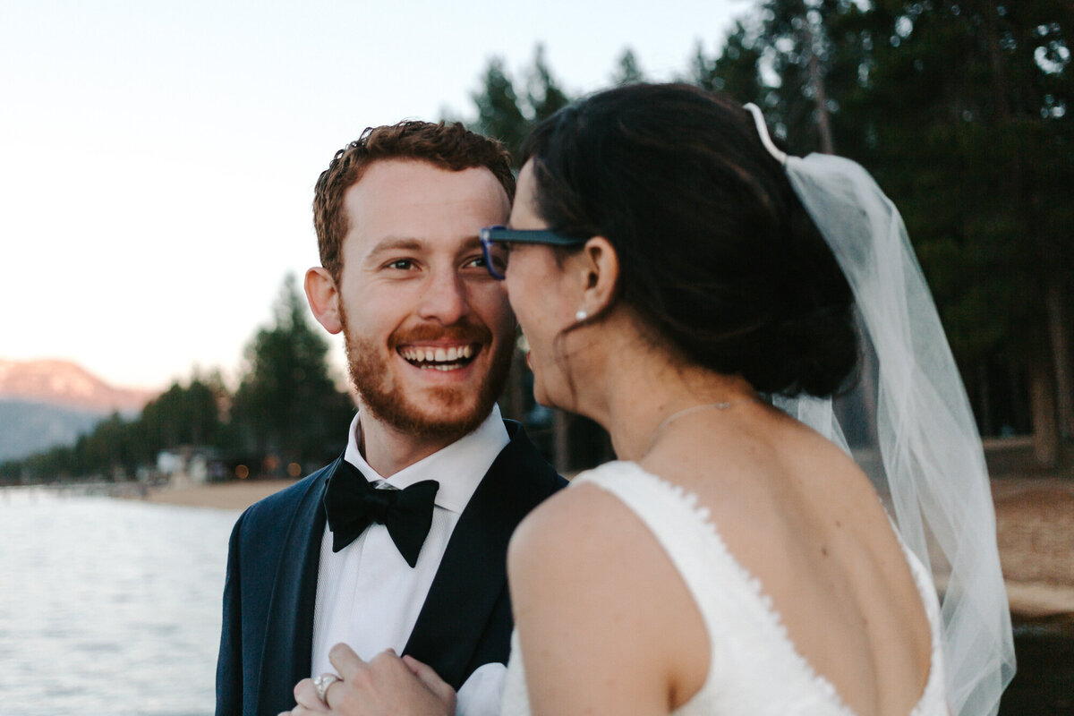 A colorful portrait of Rebecca and Damiano on their wedding day at Camp Richardson in Lake Tahoe, California. The bride and groom are on a dock in the lake with the pine trees and mountains behind them, lit up by the sunset. The groom is facing us with a wide smile on his face while the bride snuggles in. They are both laughing and enjoying the moment. Wedding photography by Stacie McChesney/Vitae Weddings.