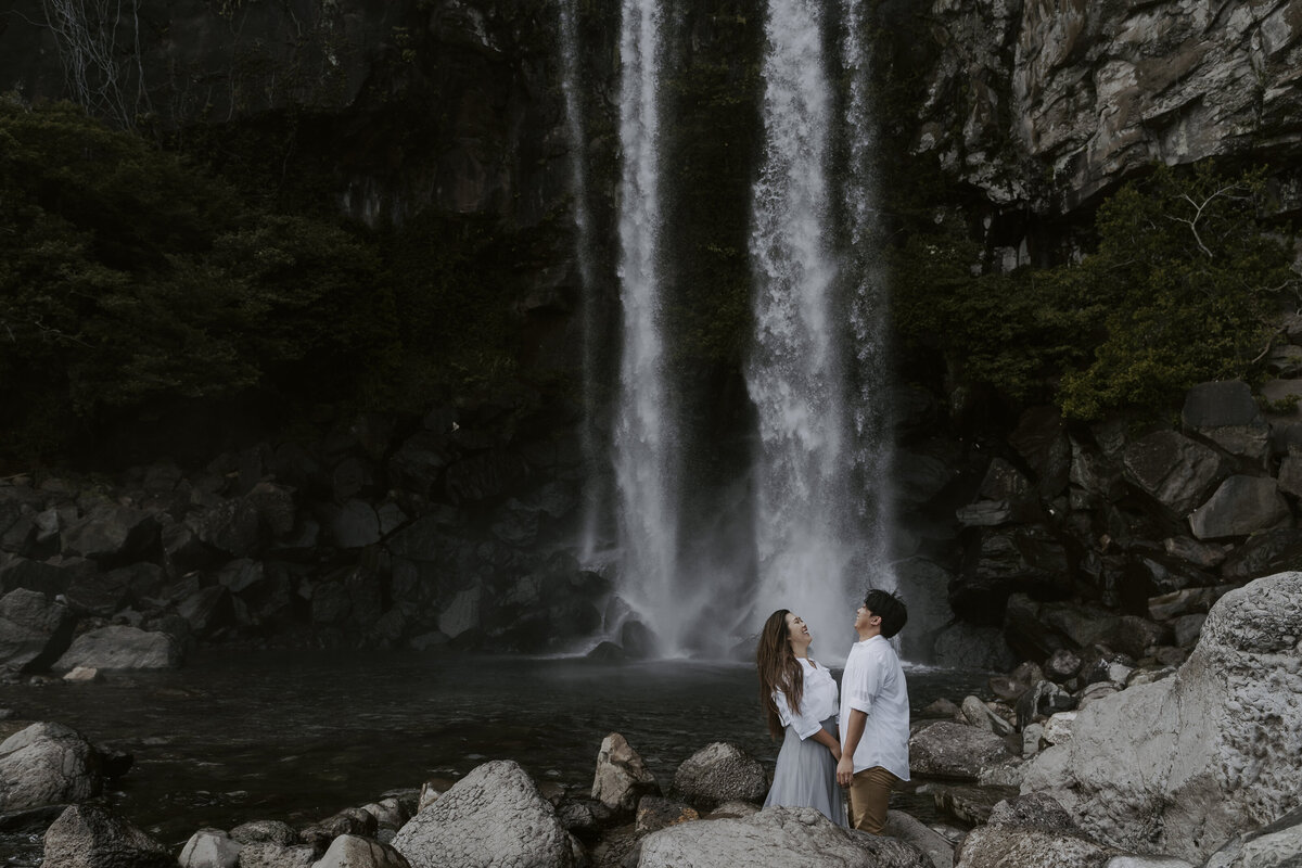 the couple holding hands and laughing in front at jeongbang waterfall in jeju island