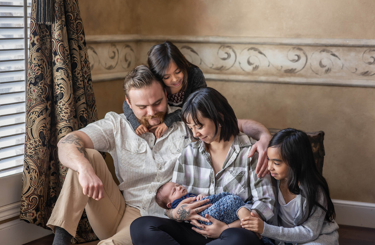 Family newborn session with dad, mom, sisters, and newborn brother .  Everyone is looking at the newborn brother while smiling