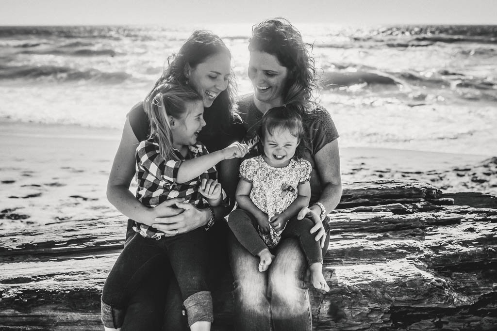 Same sex family portrait on beach with everyone smiling.  Two moms hold daughters. Big sister tickles younger sister with feather.