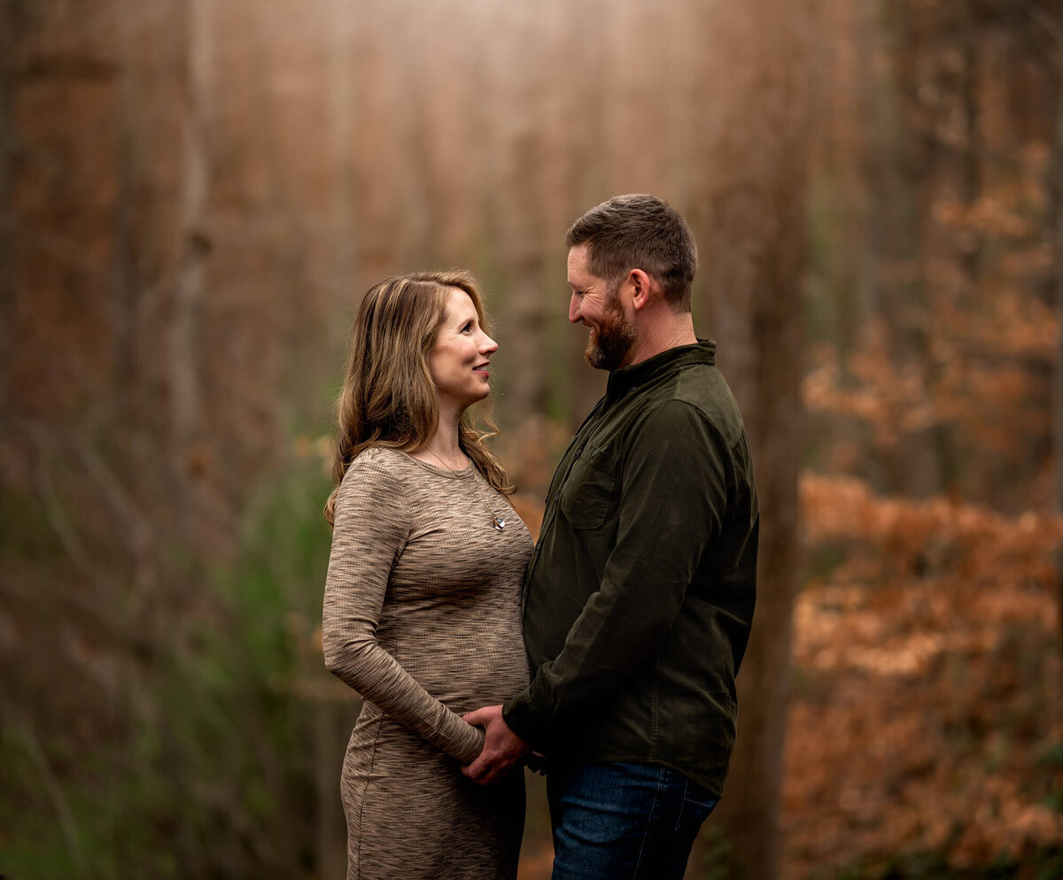 An expecting couple standing in the forest and  cradling the baby bump