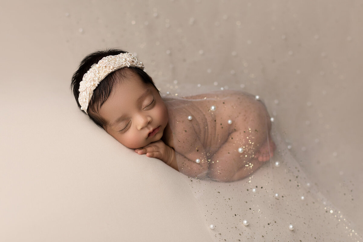 FIne art newborn photo. Side profile image of bare baby girl sleeping on her belly with her head turned toward the camera.. She is draped in a pearl-embellished sheer organza fabric and wearing a cream headband. Her hand is resting under her cheek. Captured by New Jersey's best newborn photographer, Katie Marshall.
