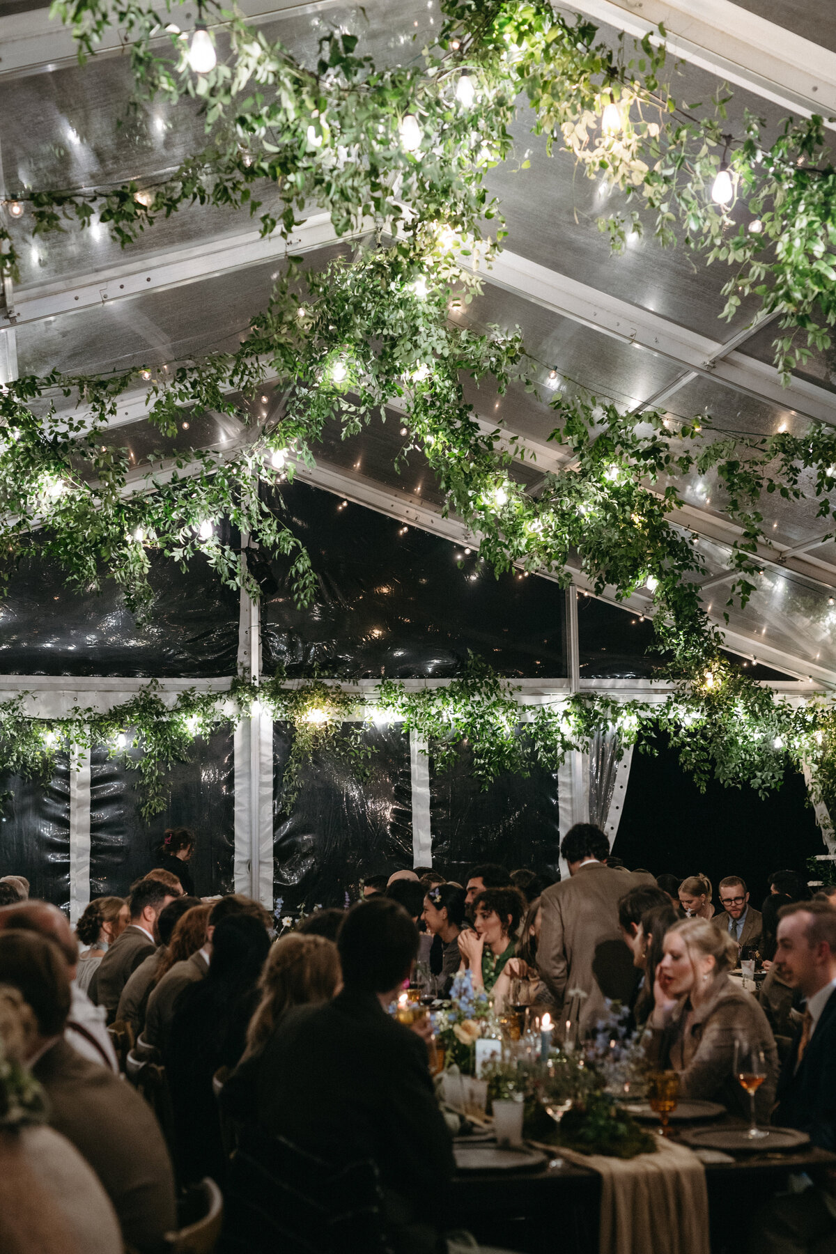 Wedding guests dining beneath a greenery-draped clear tent illuminated with twinkle lights at The Nest in Northwest Arkansas. Romantic reception atmosphere with lush garden-style floral centerpieces designed by Fleurish Floral Studio.