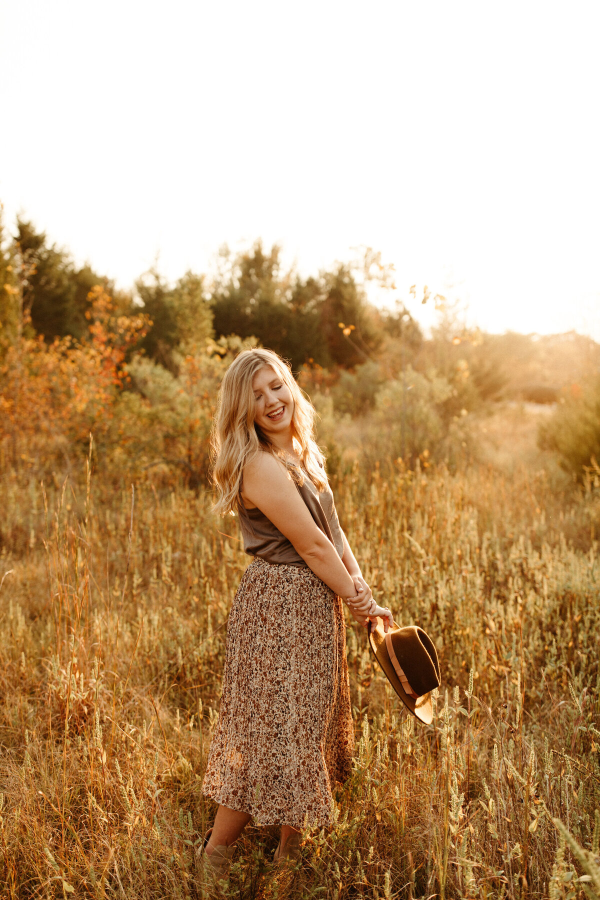High school senior in brown shirt tucked into a long floral skirt twirling in a golden field at sunset