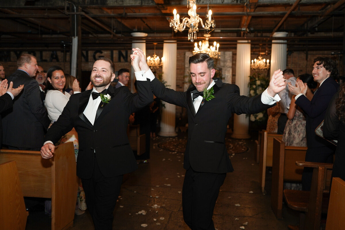 Two grooms wearing tuxedos happily raise arms and smile walking down aisle for the first time as husband and husband at Chicago wedding.