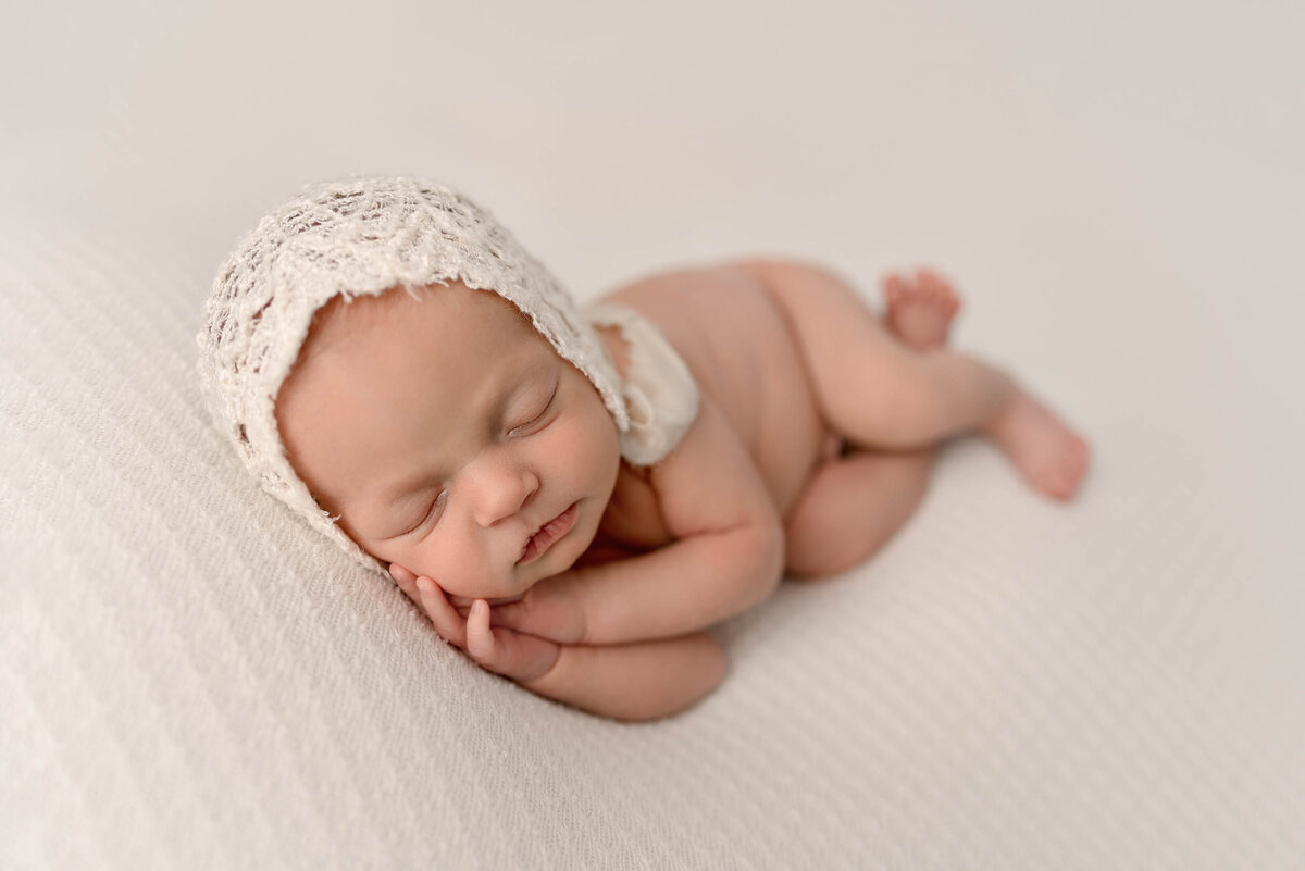 Newborn baby girl laying on her side with hands under cheek wearing a white bonnet
