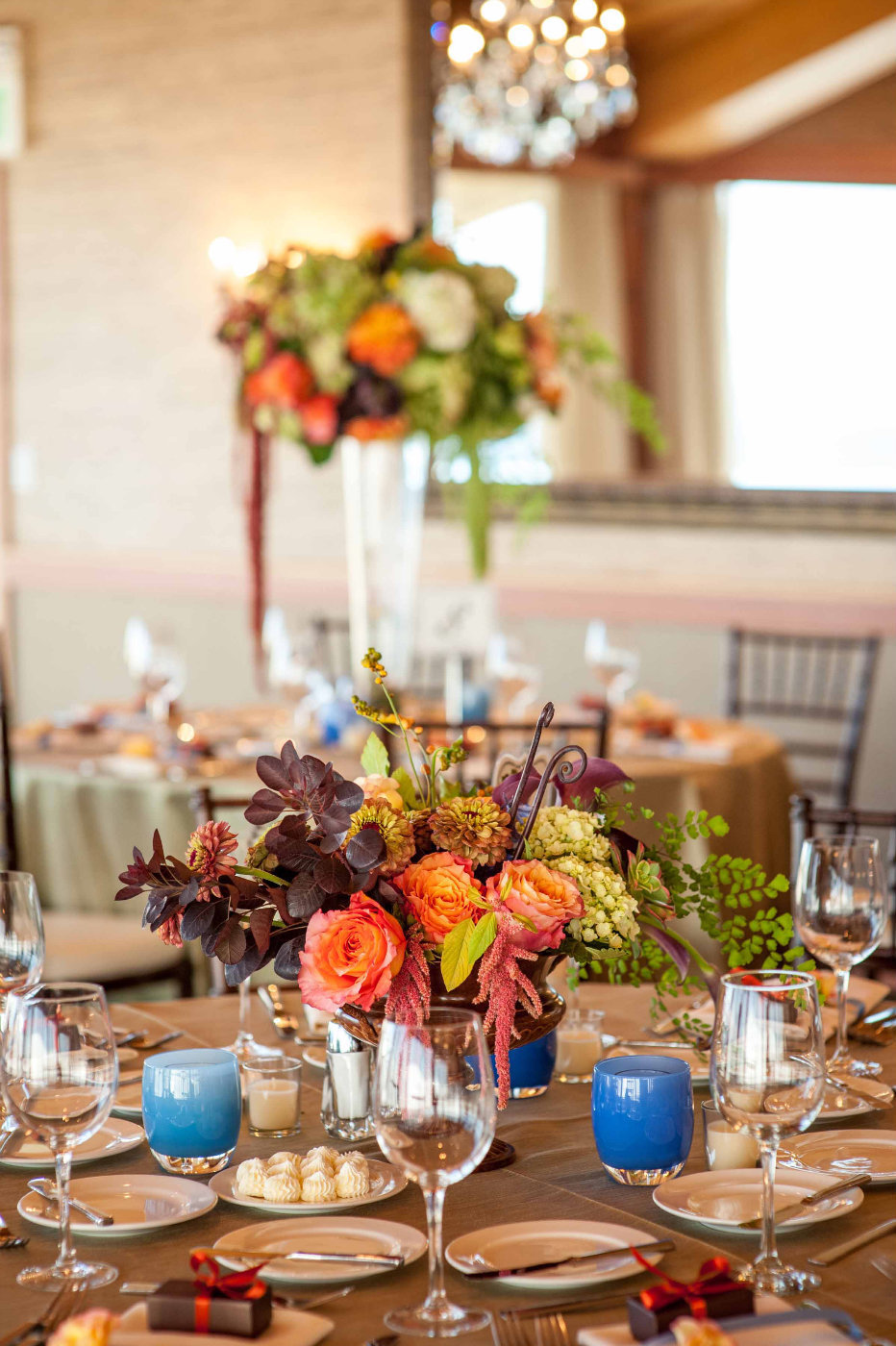 fall centerpiece of orange roses, succulents, purple calla lilies, ferns, green amaranthus, zinnia, fiddlehead fern in brown urn with blue glassybaby candle with matching tall centerpiece in back