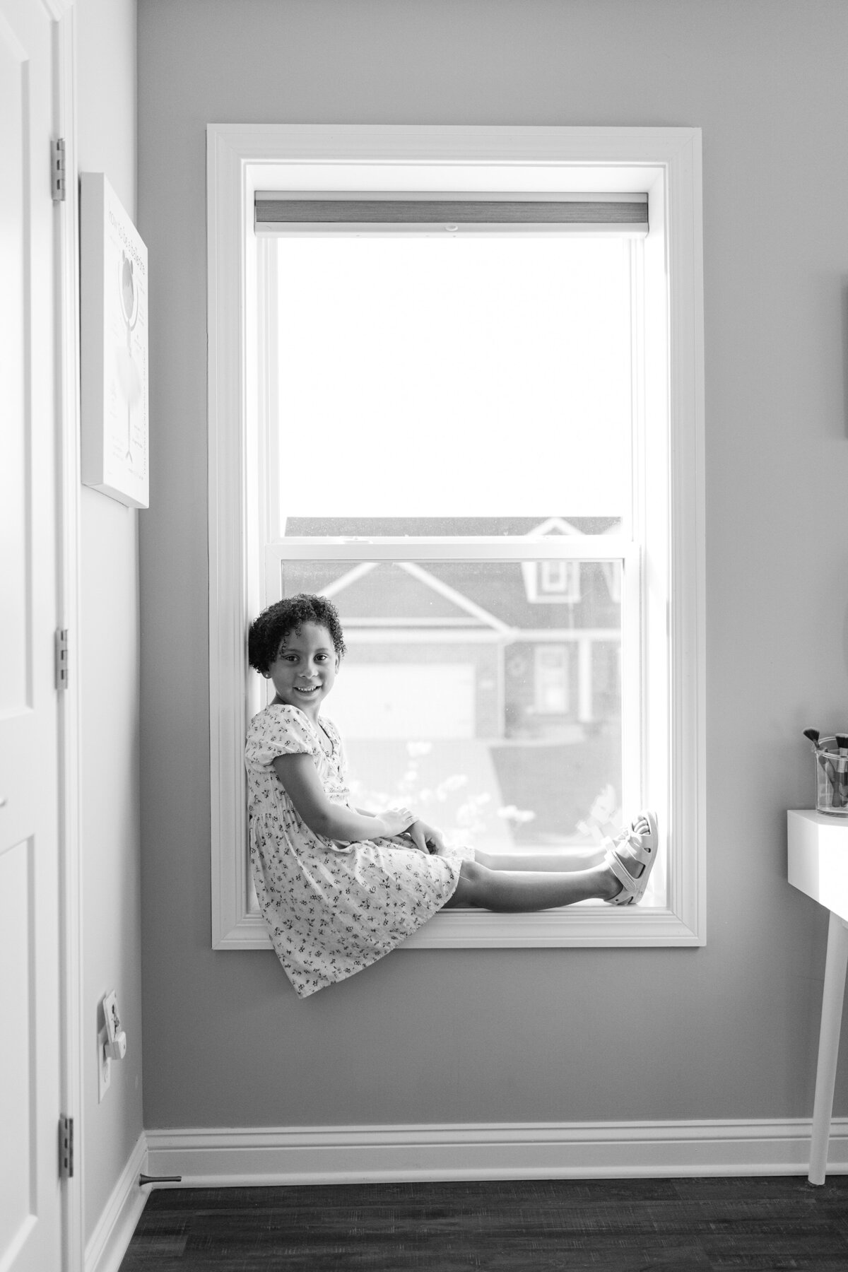 A black and white image of little girl sitting in window.