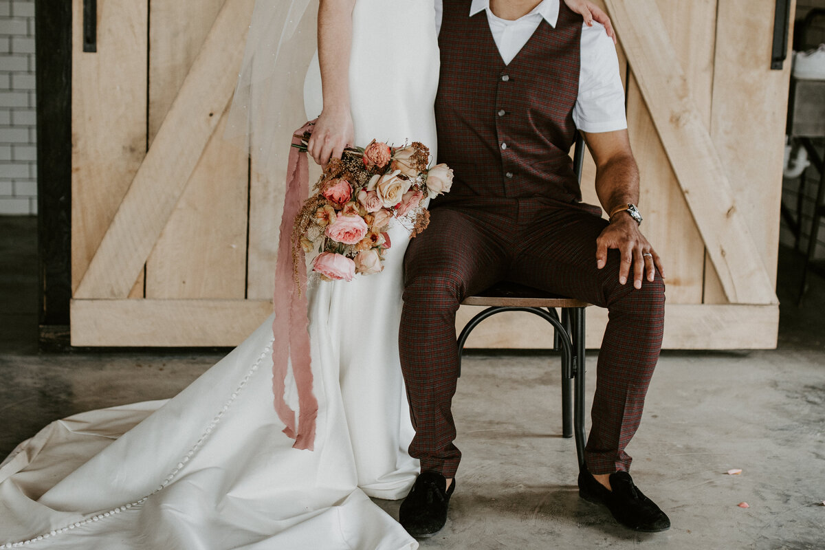 A standing bride wearing a white wedding gown holds a bouquet of blush flowers wraps her arm around groom wearing brown.