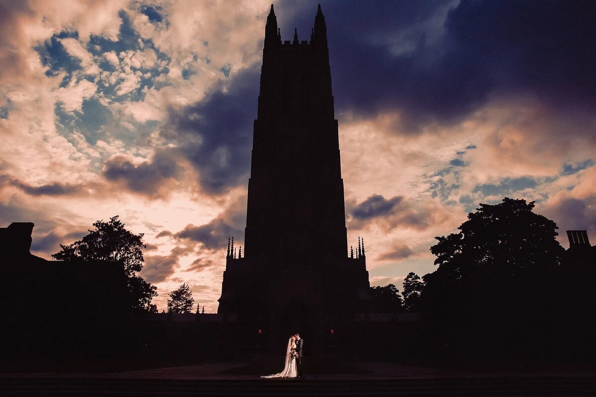 Silhouette of a bride and groom in front of Duke Chapel at sunset, with the sky painted in hues of blue and orange