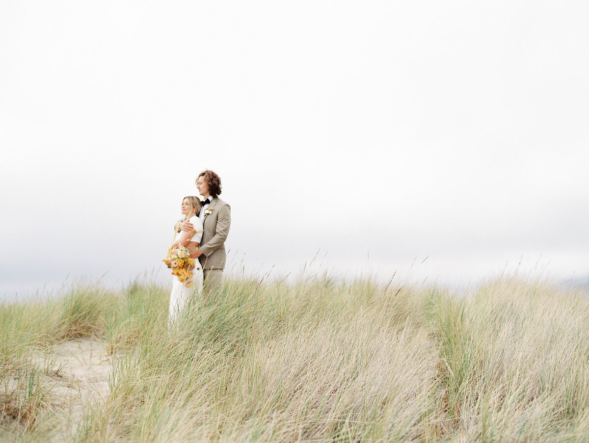 a bride leaning back into her groom on a hill of seagrass in cannon beach oregon