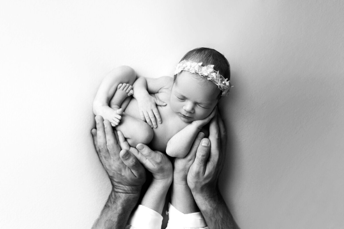 Black and white fine art newborn photo. Baby girl is sleeping with her feet folded into a ball. One hand resting on her chest, the other on her cheek. Under her, mom and dad's hands are cradling baby. Captured by New Jersey's best newborn photographer Katie Marshall.