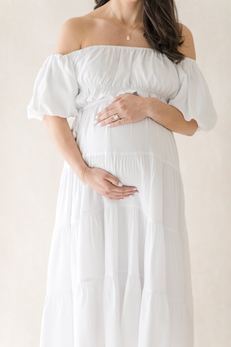 A pregnant woman wearing a white off the shoulder dress holds her stomach  during her Lawrenceville NJ Maternity Photography session