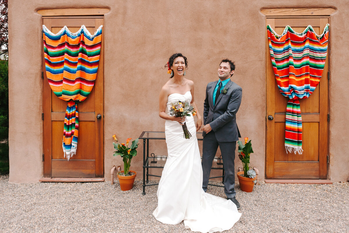 A colorful photograph of Danielle and Chris on their wedding day at Private Residence in Taos, New Mexico. The bride and groom are laughing and standing in front of stucco adobe. There are two wooden doors on either side of them. Both are drapped in a colorful, striped cloth, cinched at the center. Wedding photography by Stacie McChesney/Vitae Weddings.
