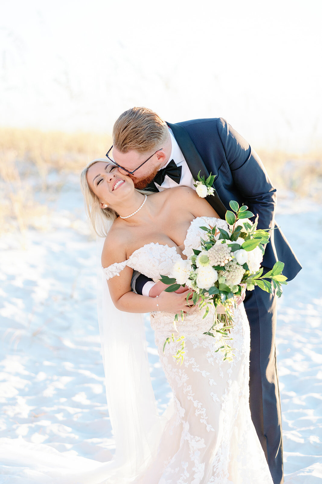 Romantic image of the bride and groom on the beach outside the Don Cesar Hotel in St. Pete Beach, FL. The groom embraces the bride from behind, kissing her cheek, during their December wedding.