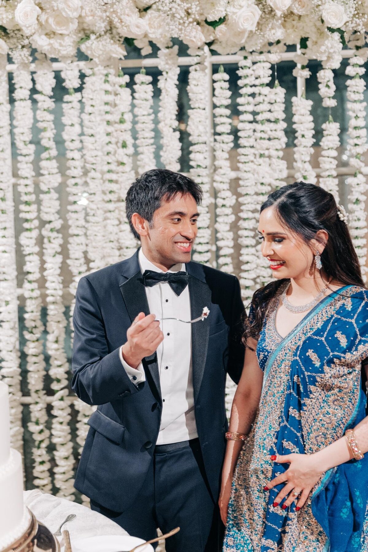 A bride in a blue saree and a groom in a tuxedo smile at each other at their wedding, standing in front of a floral backdrop.