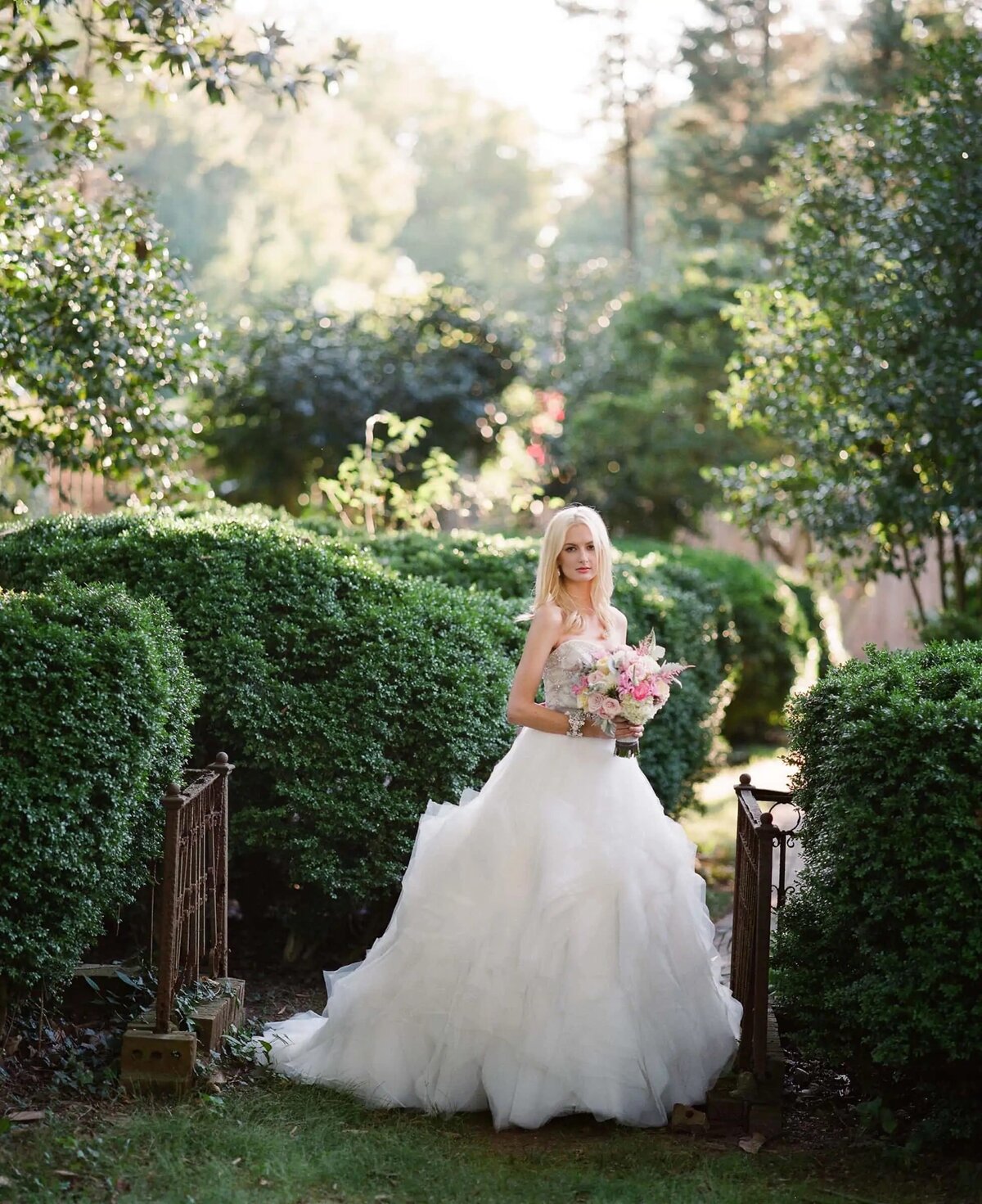 A bride stands in a lush garden, her voluminous gown contrasted with the structured greenery around her, holding a bouquet, poised and elegant.