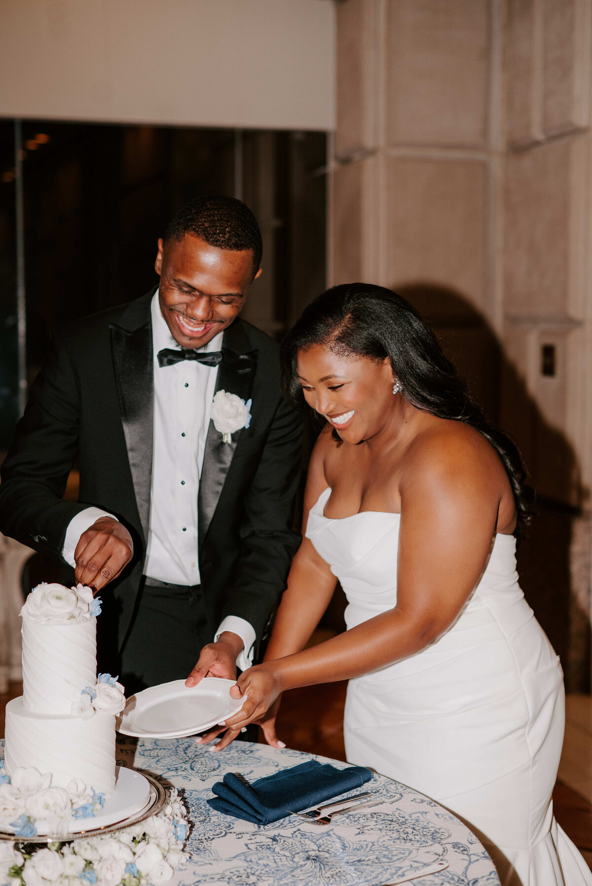 A joyful couple in formal attire cuts a white, decorated wedding cake. They are smiling and focused, creating a warm and celebratory atmosphere.