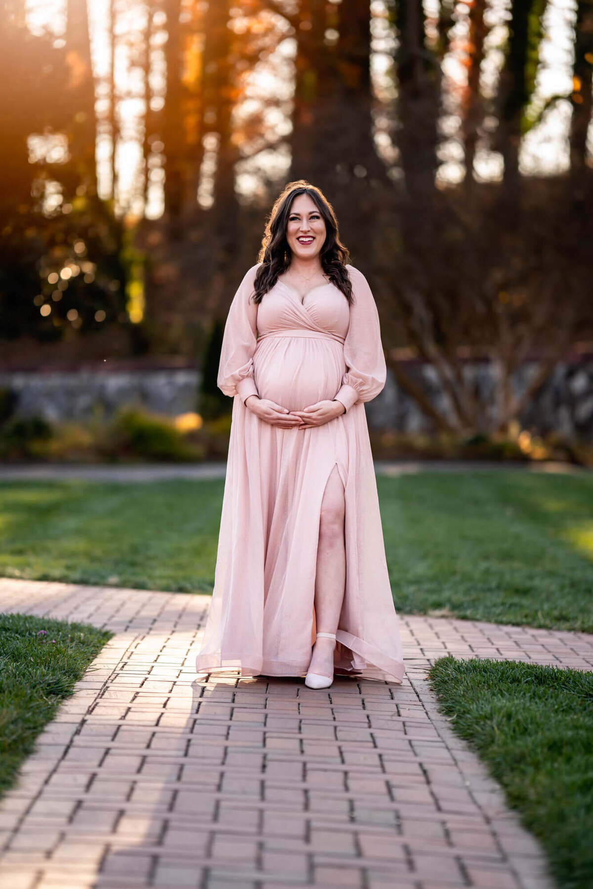 An expecting young woman in a long pink dress cradles her bump and smiles for the camera during her portrait session with an Asheville maternity Photographer