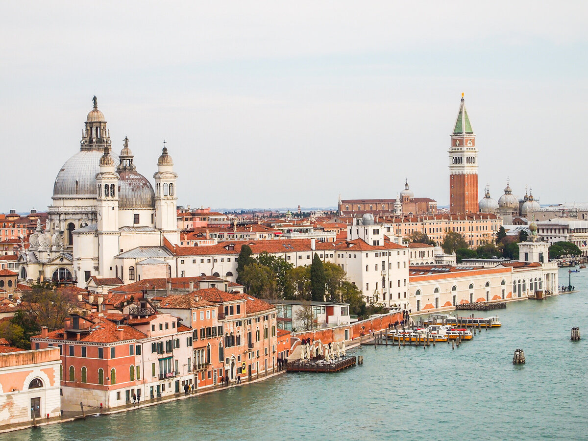 Aerial view of Venice, Italy, featuring historic buildings along a canal. Prominent landmarks include a large domed church and a tall bell tower. Boats are moored along the waterfront, with more buildings stretching into the background.