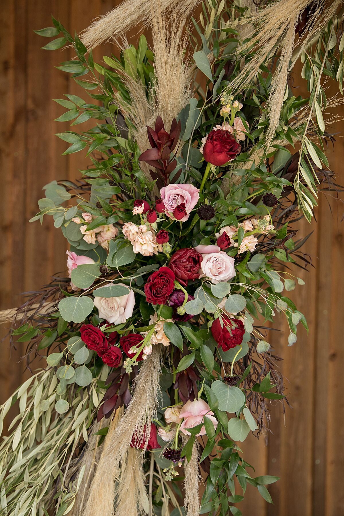 Boho ceremony arbor flowers with eucalyptus, olive branches, blush, pink and ivory roses, pampas grass and burgundy roses and peonies.