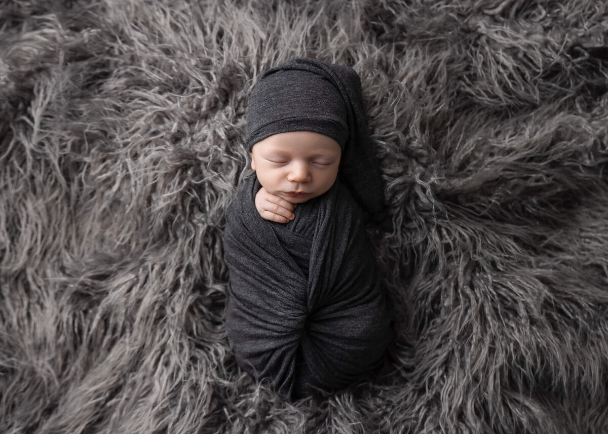 newborn baby wrapped in grey fabric wearing a grey sleepy cap on a grey shaggy rug