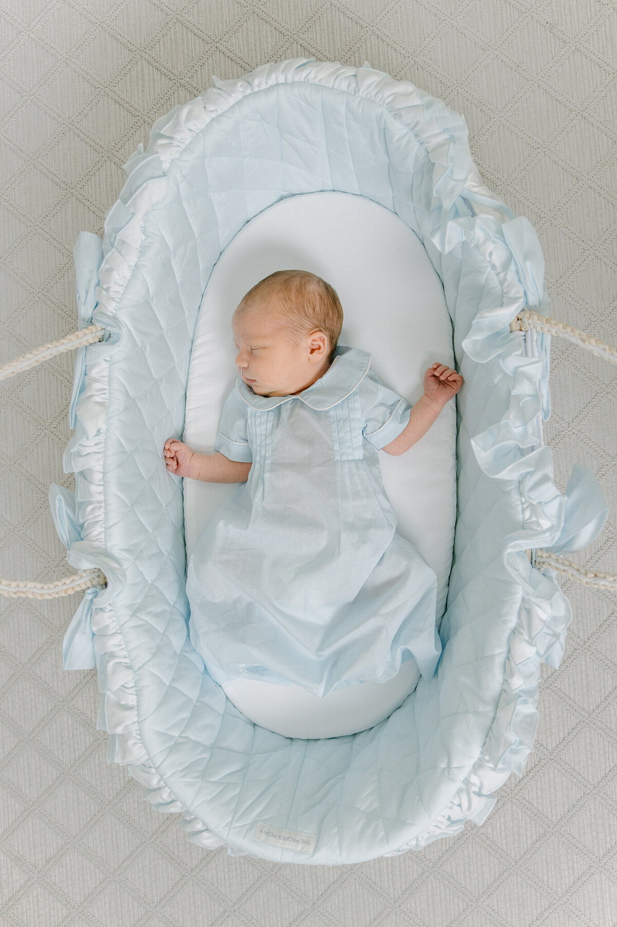 Newborn sleeping in a blue ruffle moses basket wearing a blue newborn gown.