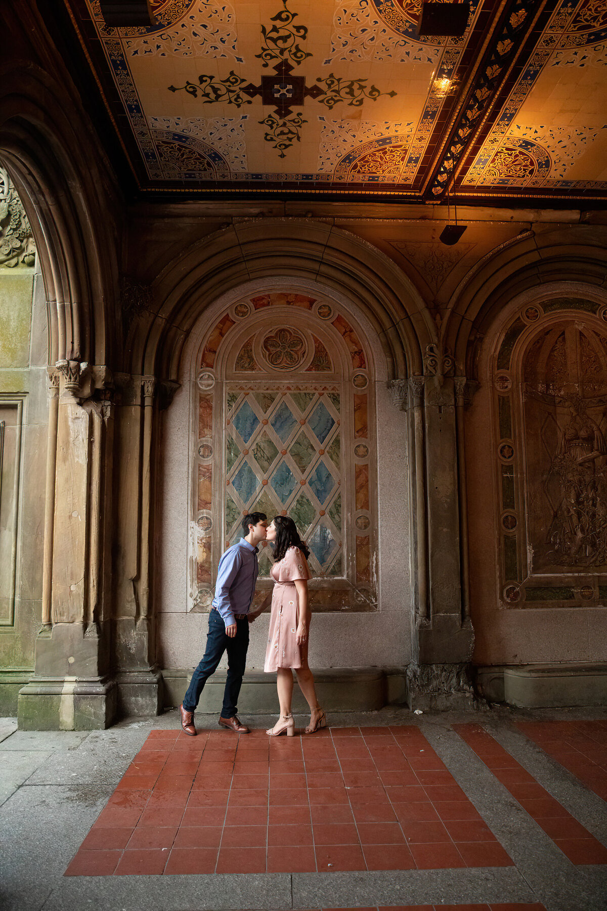 A man in a blue dress shirt and jeans kisses a woman in a pink dress by the historic mosaics and arches in Central Park Bethesda Terrance New York wedding photographer