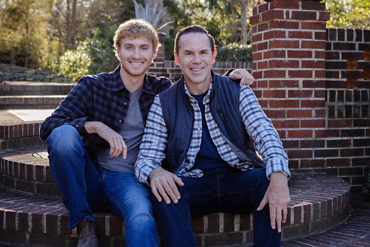 Proud autumn portrait of father with college senior seated together on brick terrace steps at UNCC Botanical Garden Charlotte NC