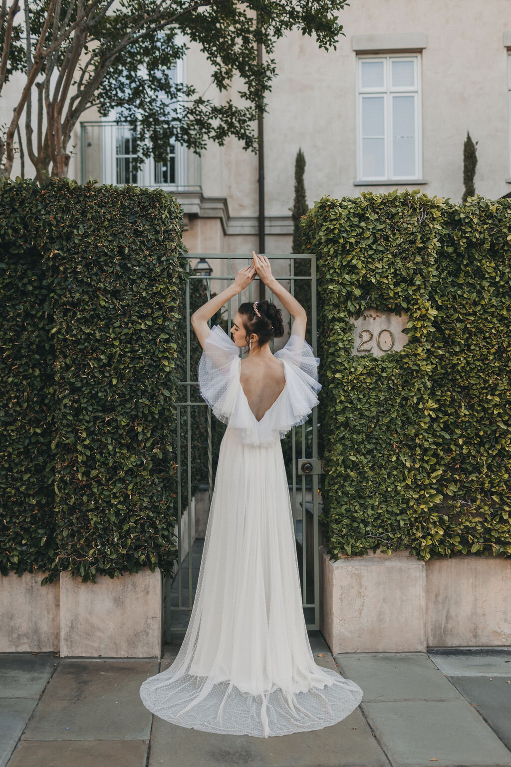 The Mai bridal style is modeled in front of a greenery wall on the Charleston peninsula. the model stands with her back to the camera and her arms above her head, which fans out the flutter sleeves into wings.