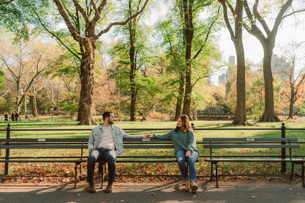 man and woman sitting on opposite sides of a park bench looking at each other while they reach for each other