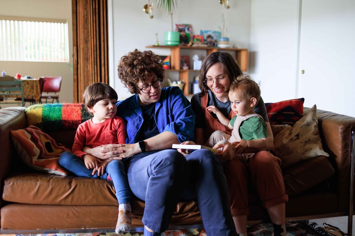 Two moms and their young kids read together on the couch during an at home family photography session.
