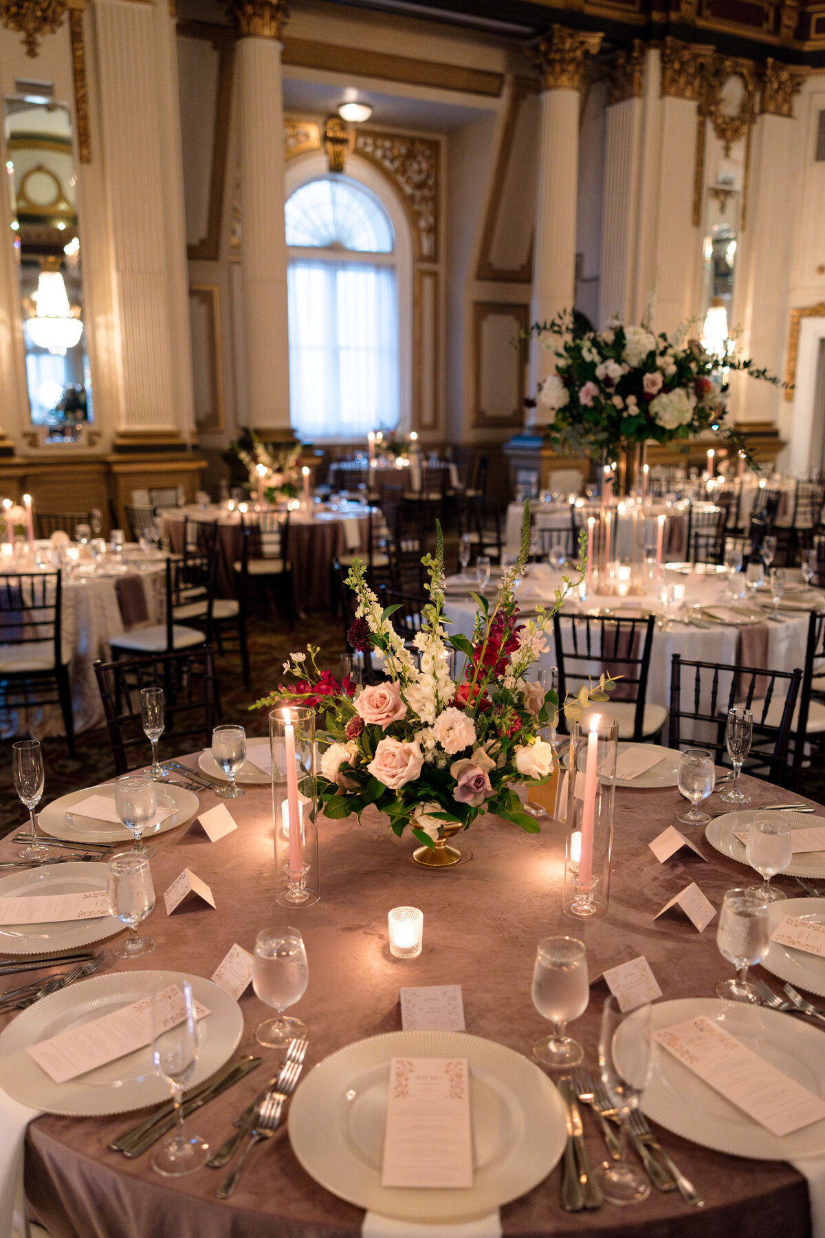 Elegant ballroom with round tables set for an event. Tables feature floral centerpieces with roses and candles, surrounded by silverware and menus. Tall windows with drapes and ornate pillars decorate the room, creating a sophisticated atmosphere.