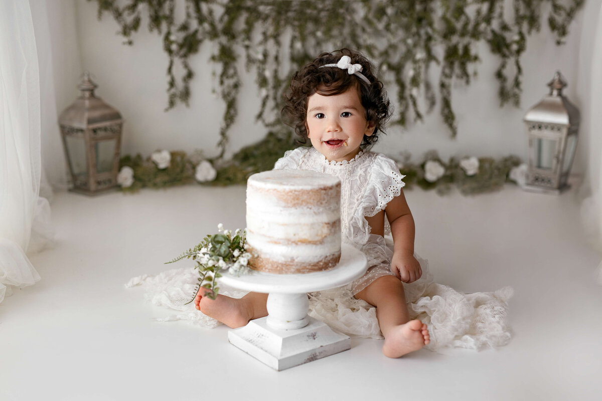 happy birthday girl sitting in front of a cake in a photography studio with greenery and flowers behind her