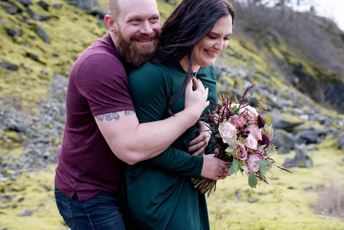 a woman holding a pretty bouquet by flowers by alana  laughs as her fiance gives her a big bear hug