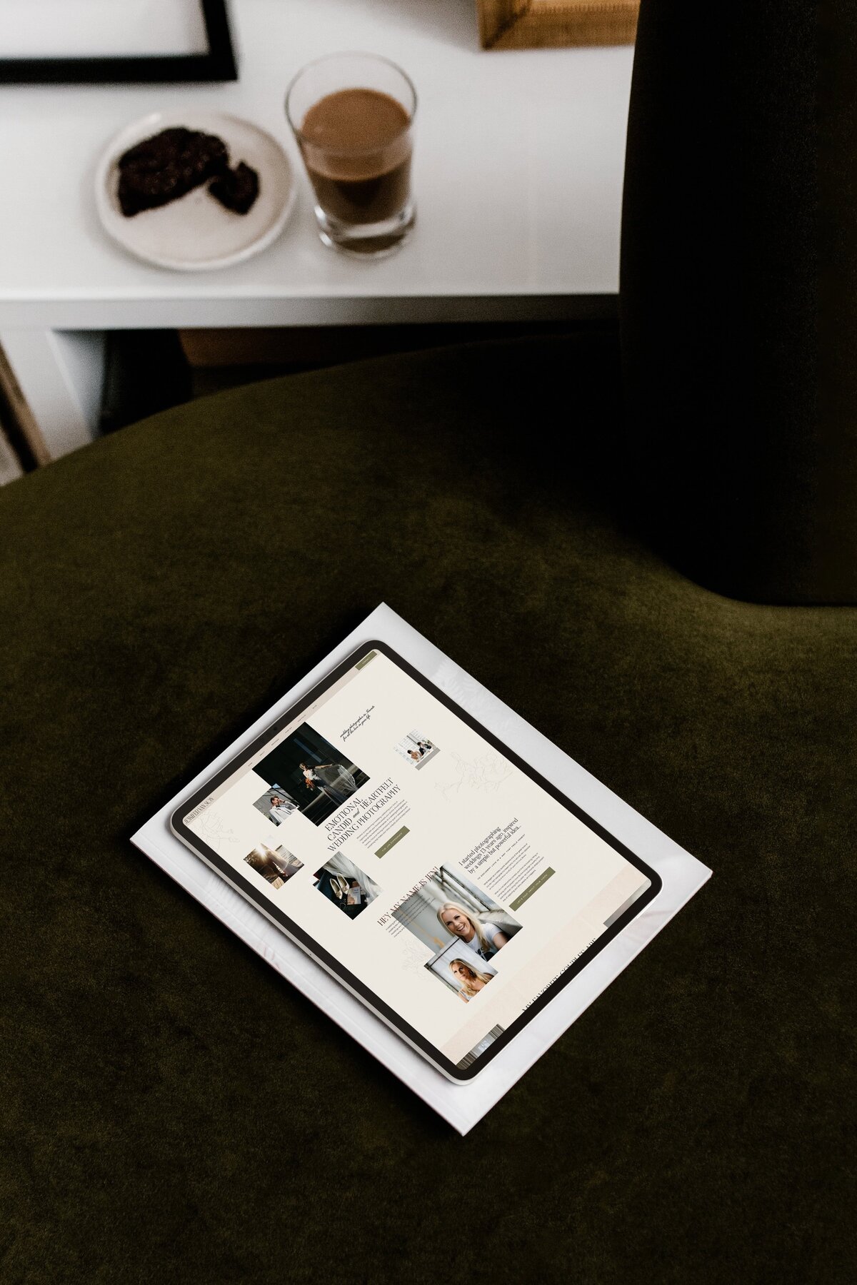 A tablet is placed on a stack of papers. The screen displays a webpage with images, text, and a light-colored layout, specializing in brand and identity services for luxury photographers. The background surface is a dark green fabric.