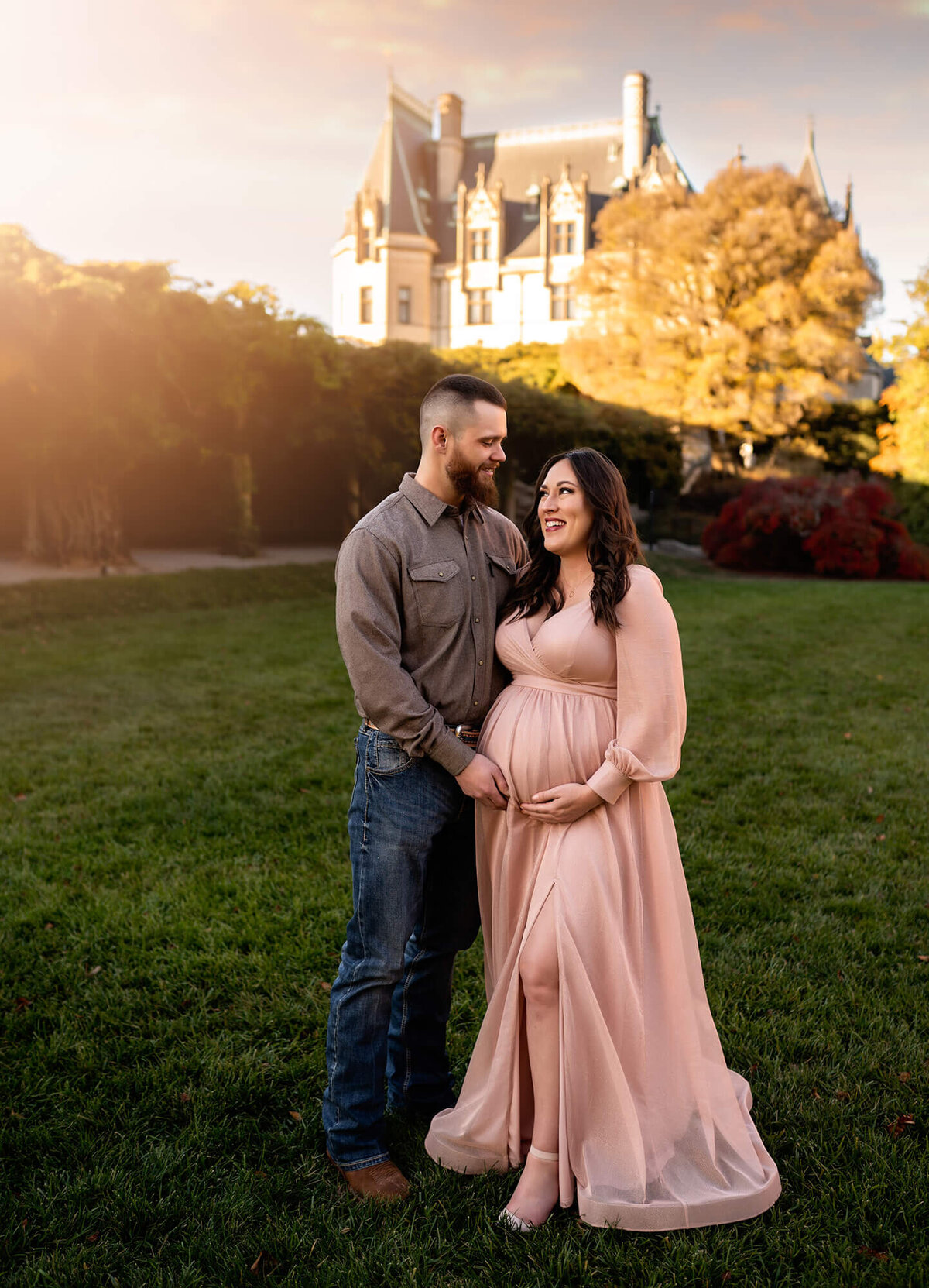 A soon to be mom and dad cuddling and smiling in front of the Biltmore Estate during their portrait session with an Asheville Maternity Photographer