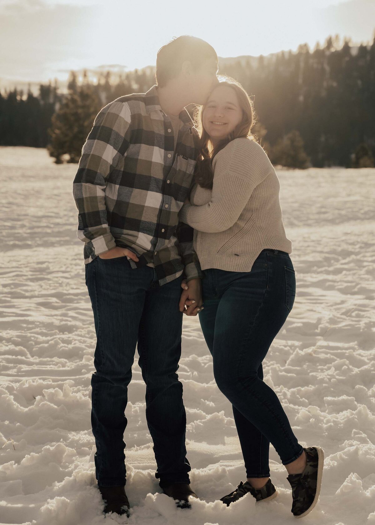Maddie Rae Photography a couple standing side by side holding hands and snuggled in. he is kissing her head and she is smiling at the camera. there are mountains and snow in the background.