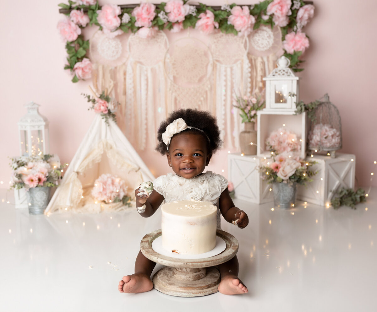 Rose Garden Cake Smash in West Palm Beach, FL. Black baby girl sitting in front of a white cake smiling at the camera with a fist full of cake. In the background, there are macrame wall hangings and roses.