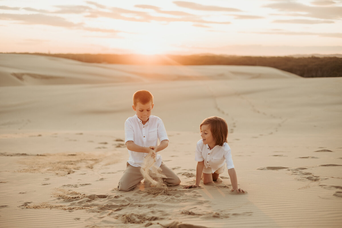 Two beautiful boys playing in the sand dune together as the sun sets behind them.